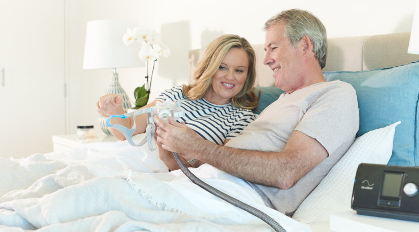 Two people sitting up in bed, reviewing a ResMed AirFit F30 CPAP mask together, with ResMed machine visible on the bedside table.