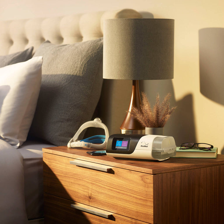 ResMed AirSense 11 CPAP Machine sitting on the nightstand next to a bed. There is a ResMed Mask behind it.