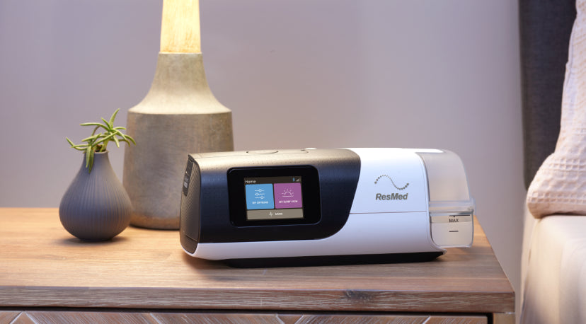 ResMed AirSense 11 CPAP machine on a wooden bedside table in a softly lit bedroom setting.