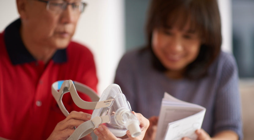 Two people examining a ResMed CPAP full face mask together, one holding the mask and the other reviewing product documentation.