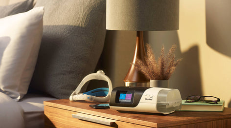 ResMed AirSense 11 CPAP Machine sitting on the nightstand next to a bed. There is a ResMed Mask behind it.