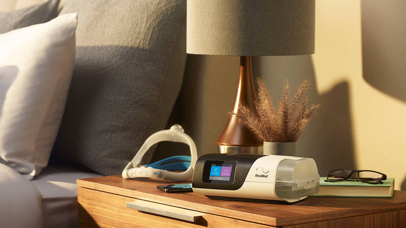 ResMed AirSense 11 CPAP Machine sitting on the nightstand next to a bed. There is a ResMed Mask behind it.